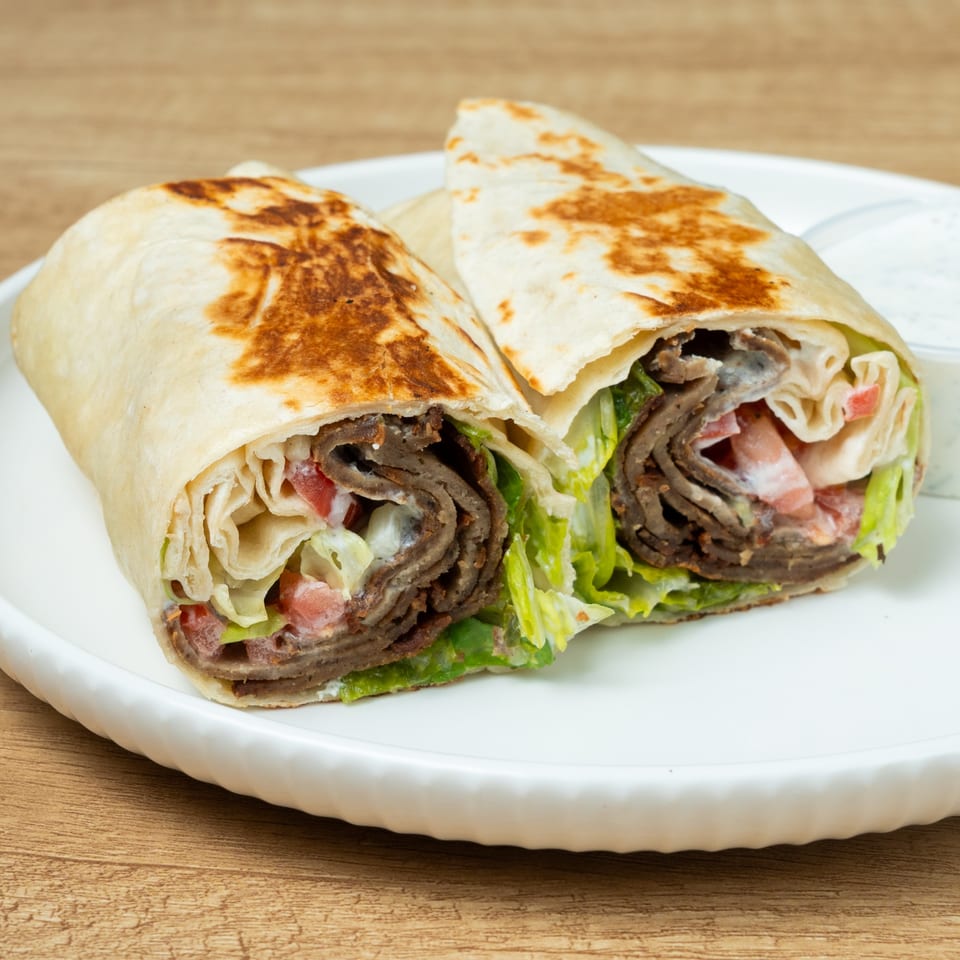 Two halves of a gyro wrap filled with meat, lettuce, tomatoes, and white sauce on a plate.