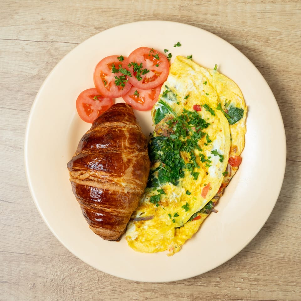 A breakfast plate with a golden croissant, a fluffy omelet with herbs, and sliced tomatoes.