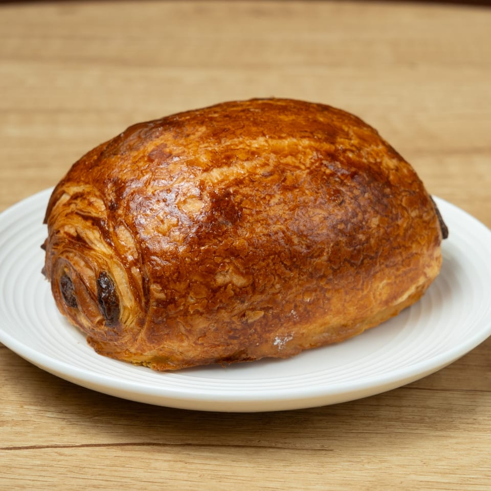 A golden-brown pain au chocolat pastry on a white plate, against a wooden background.
