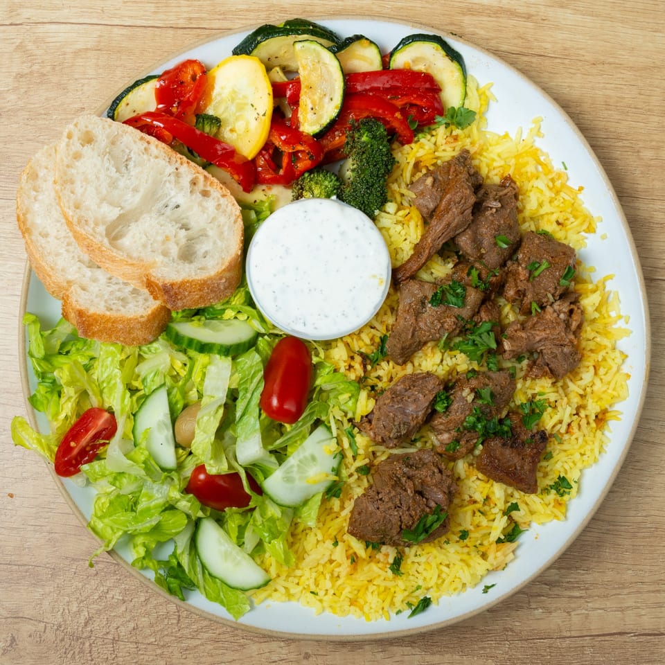 A plate of beef and yellow rice, with a side salad, grilled vegetables, bread, and a white dipping sauce.